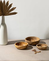 Three natural olive wood nesting bowls on a table, with a white vase and a dried palm leaf in the background.
